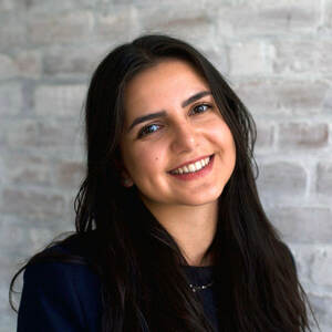 A woman with dark hair and olive complexion is wearing a dark business suit and smiling for a portrait in front of a brick wall.
